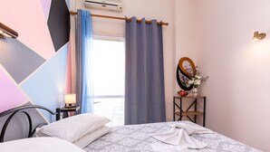 A hotel room with a bed, pillows, a bedside table with a lamp, a mirror, and a small shelf with a vase of flowers. - Riviera Seaside Hotel by Estia (Corfu)
