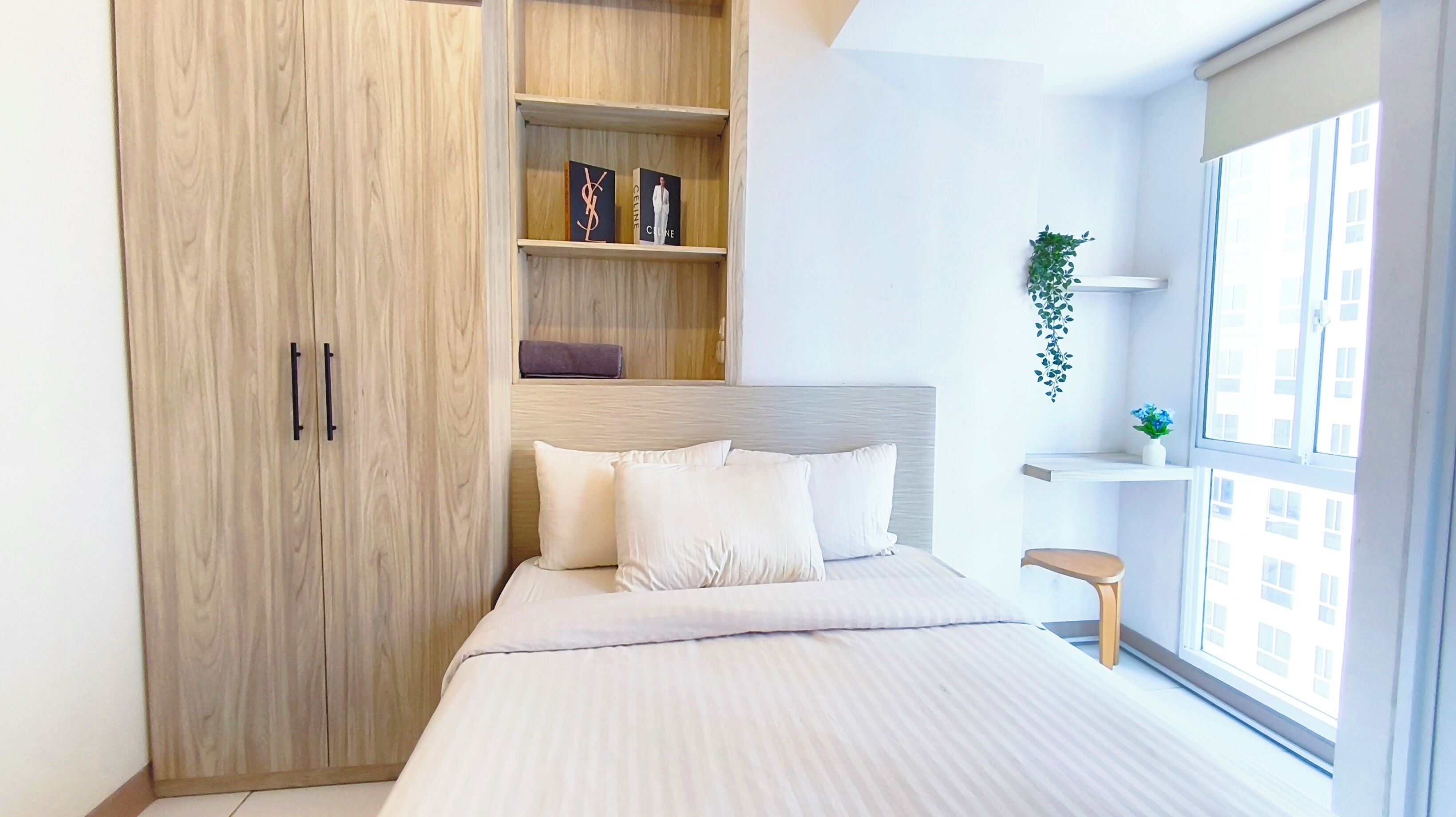 A modern bedroom with a large wardrobe, a bed with white bedding, a small bedside table, and a window with a view of buildings.