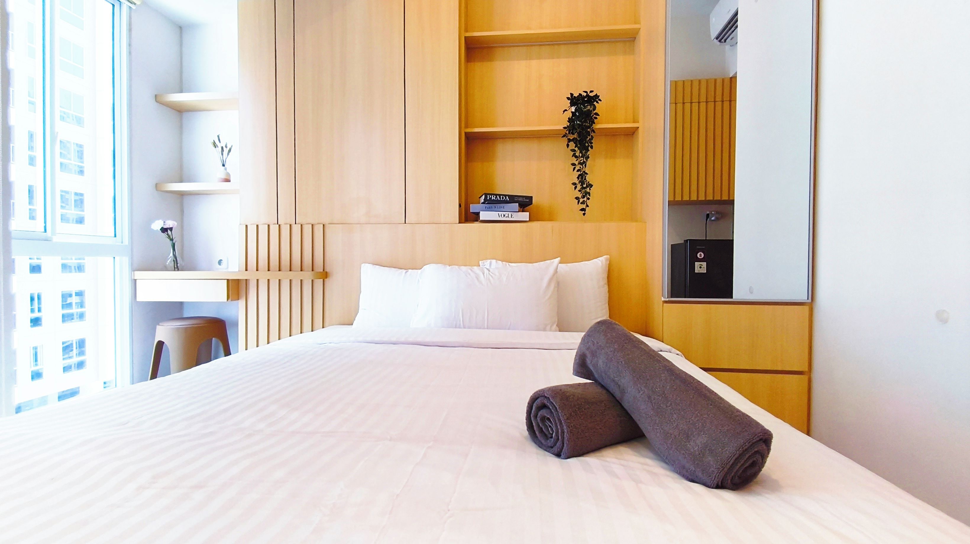 A hotel room with a bed, a desk, a chair, a bookshelf with magazines, and a window with a view of buildings.