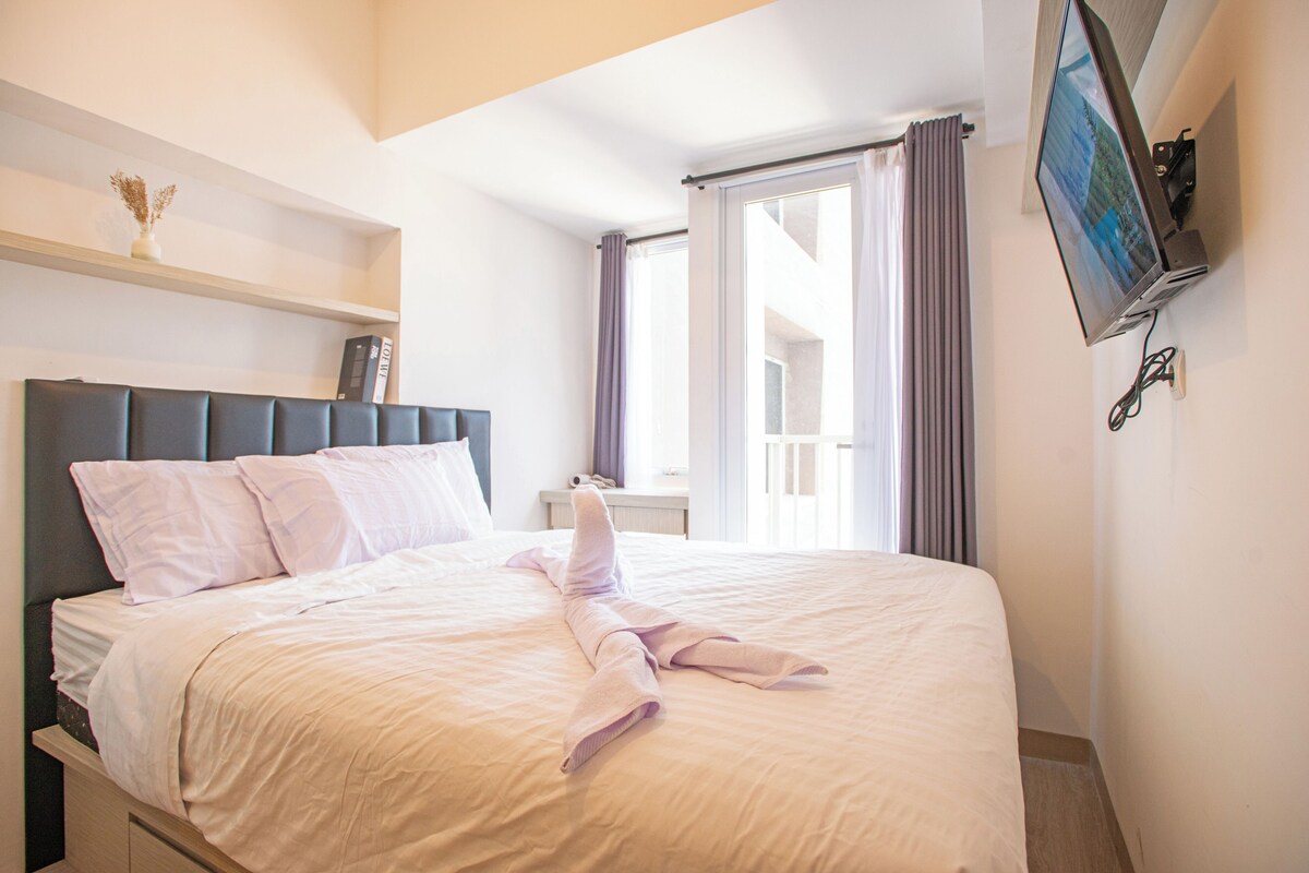 Una cama bien hecha con una toalla doblada, una mesita de noche con una lámpara y un televisor montado en la pared.