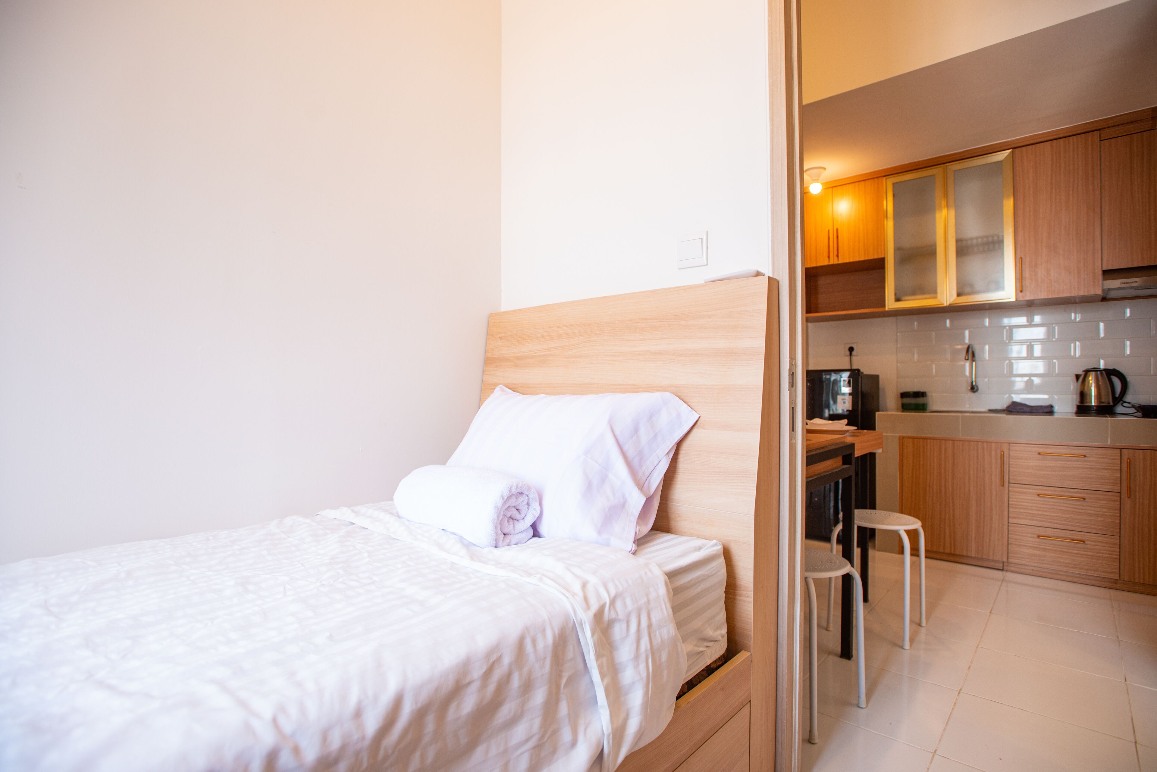 Une chambre avec une tête de lit en bois, un lit recouvert de draps blancs, un petit tabouret et un coin cuisine doté de meubles en bois et d’une bouilloire.