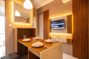 A modern hotel room with a wooden dining table set for two, a flat-screen TV mounted on the wall, and a large mirror above the table.