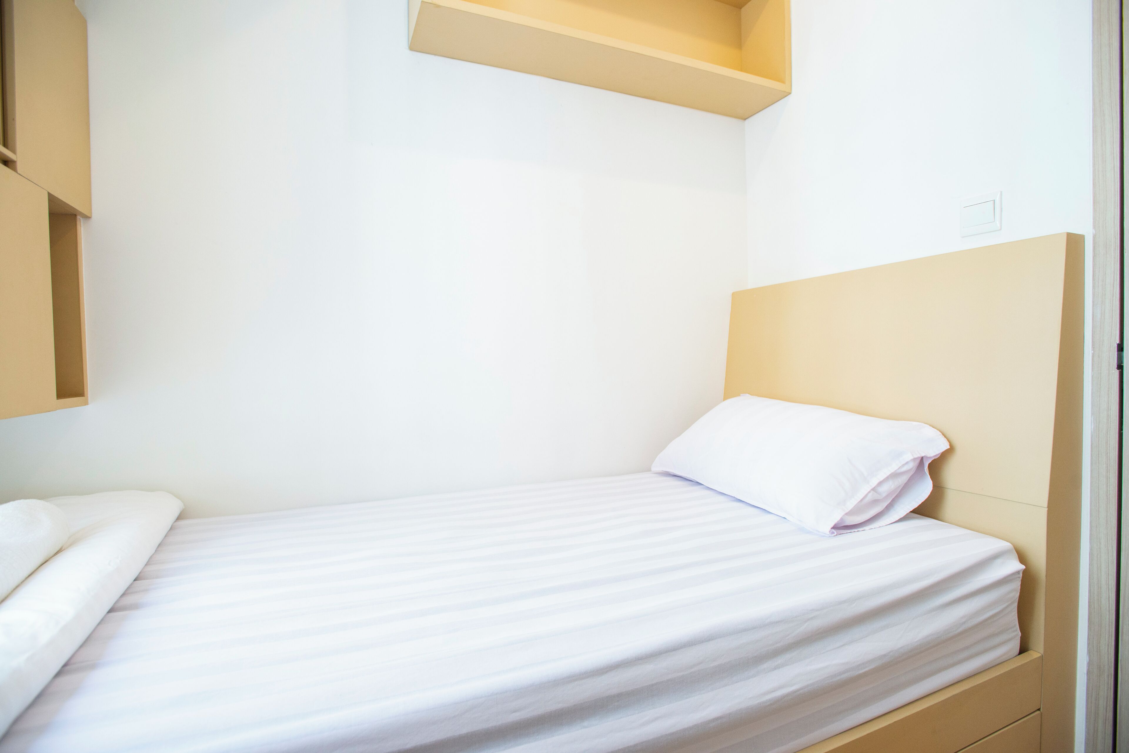 Una cama individual con colchón y almohada blancos, cabecera de madera y pequeña estantería sobre la cama.