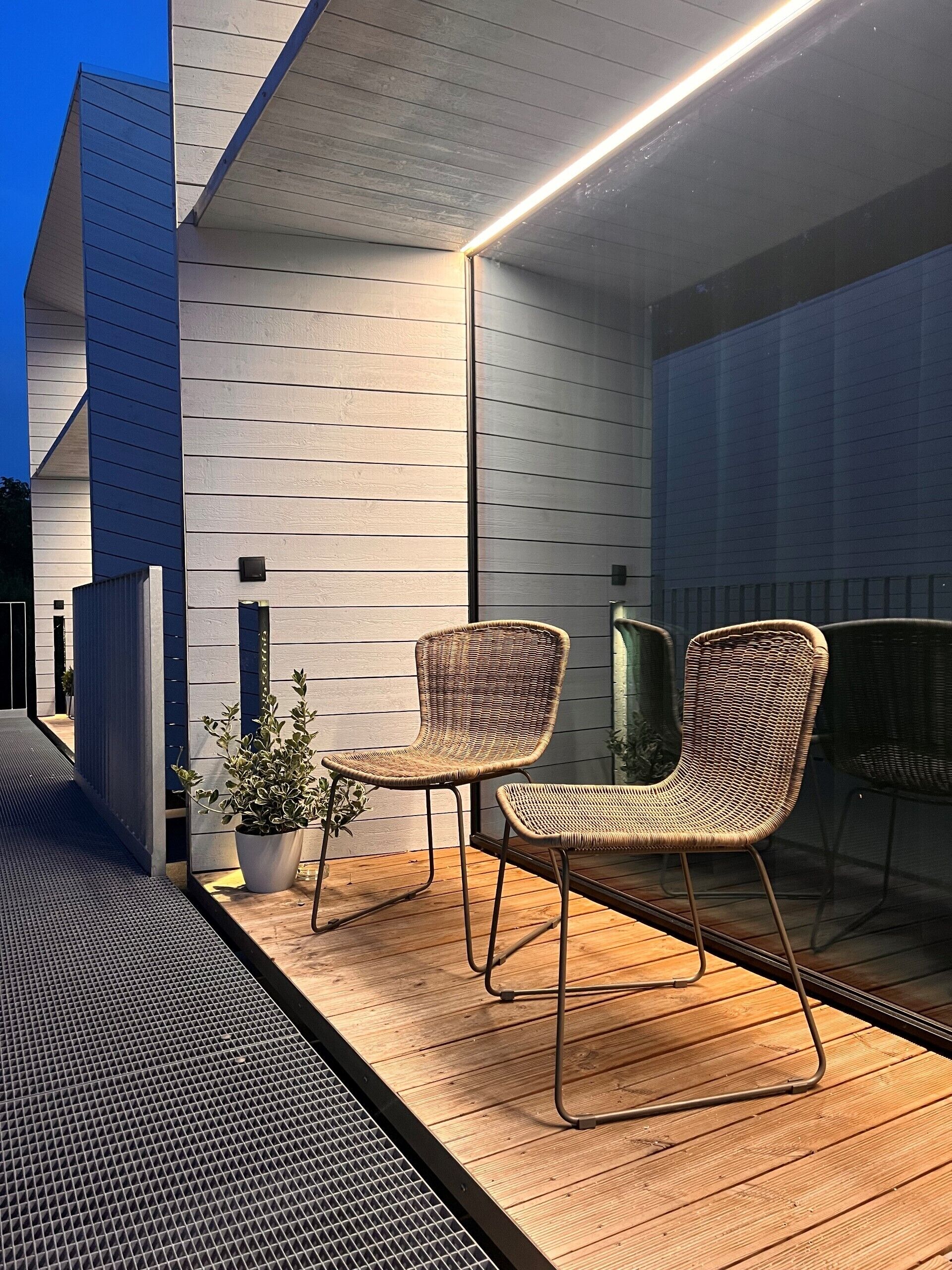 Un patio exterior con dos sillas de mimbre y una planta en maceta, junto a un edificio de diseño moderno y una puerta de vidrio.