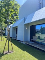 Un edificio moderno con techo inclinado, un pequeño espacio al aire libre con sillas y un árbol en primer plano.
