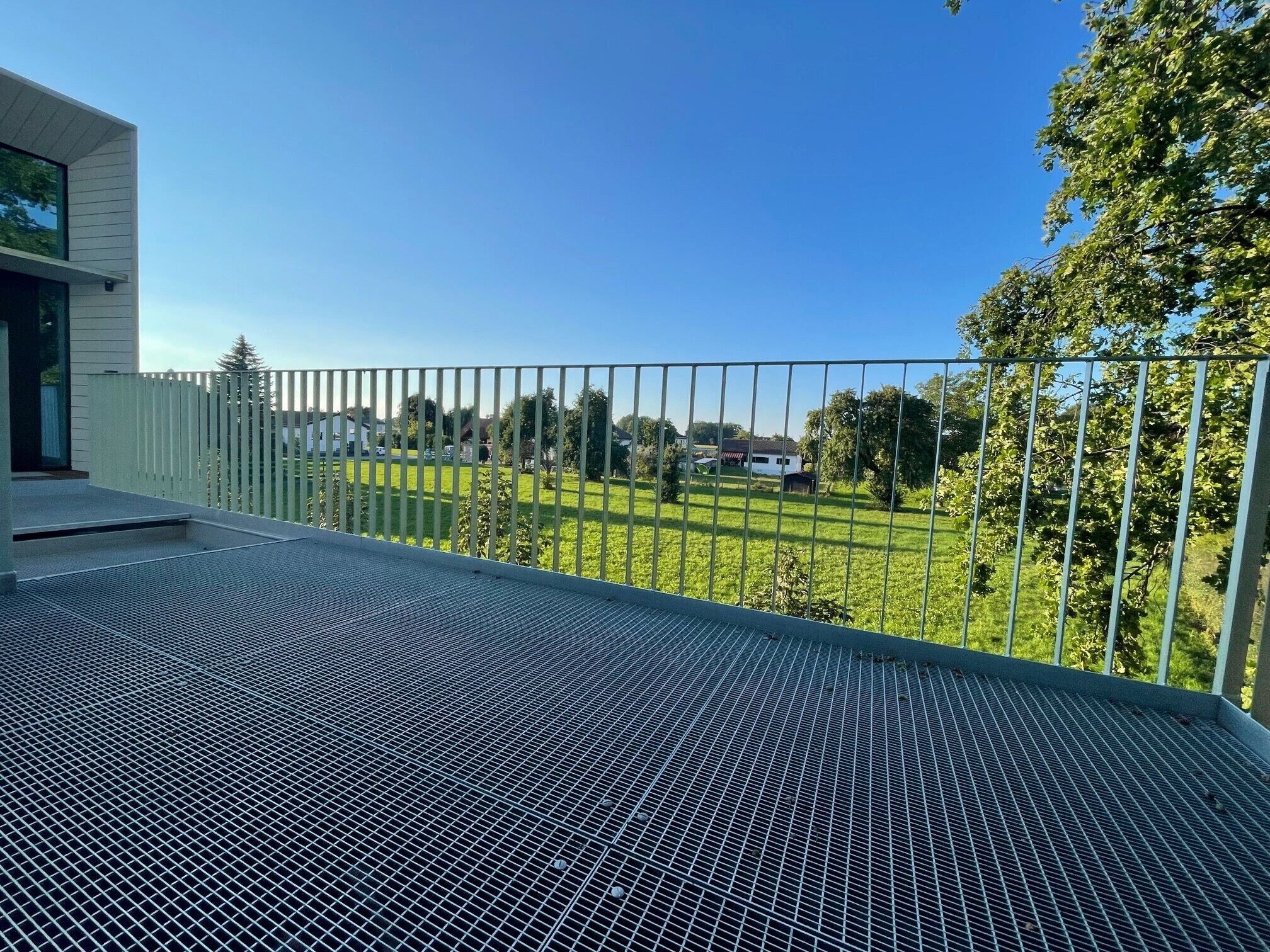 A balcony with a metal grid floor, overlooking a green lawn and a white fence.