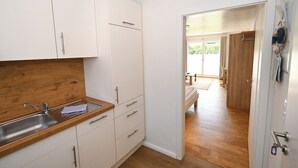 Eine Küche mit weißen Schränken, einer Holz Arbeitsplatte und einem Spülbecken. Eine Tür führt in einen Wohnbereich mit Holzboden und einem Bett.