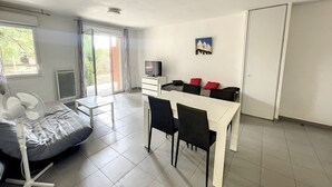 Ett kompakt bostadsutrymme med en matplats, en säng med en fläkt, en TV och en skjutdörr i glas som leder ut till uteplatsen.