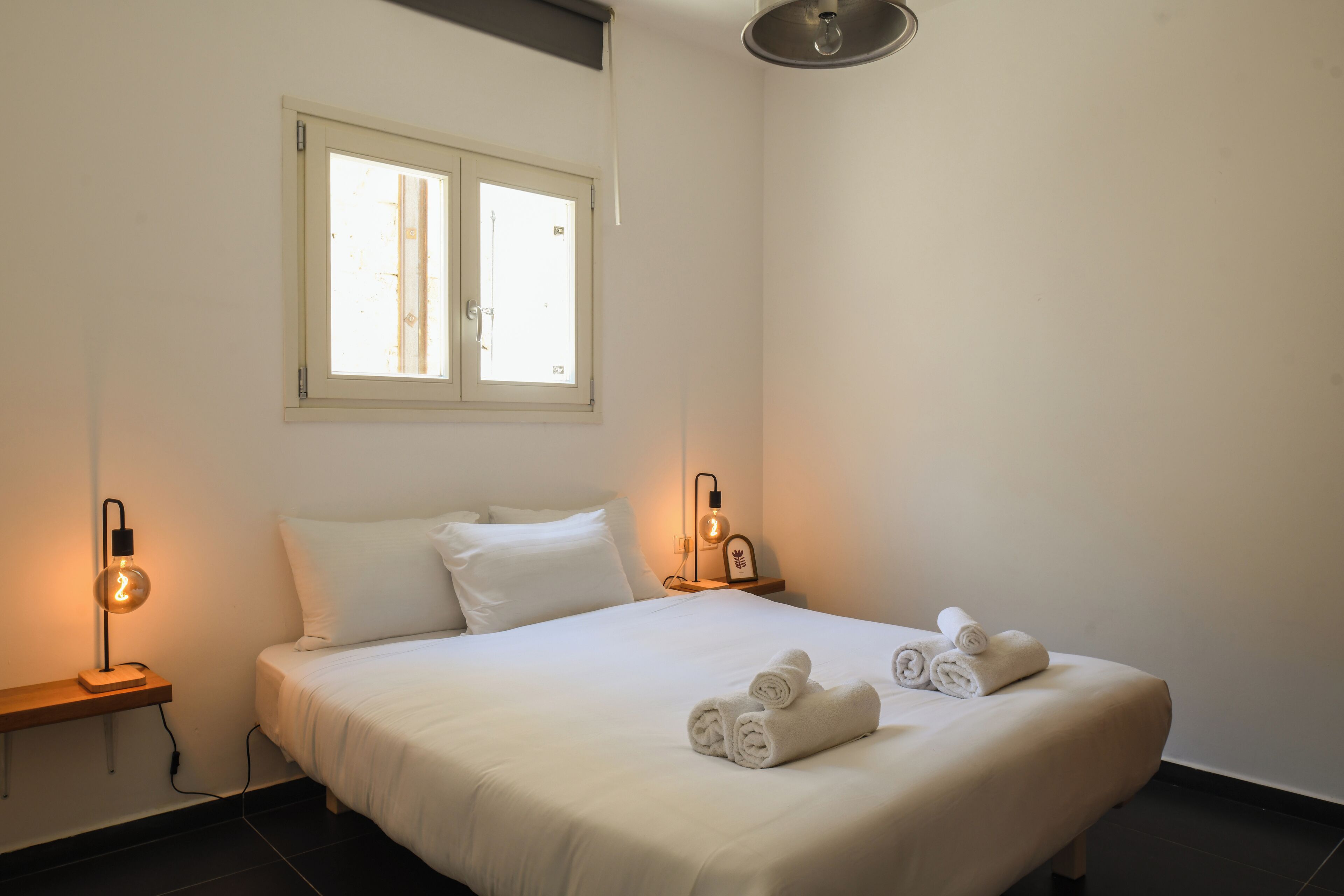 Un lit bien fait, avec du linge de lit blanc et des serviettes pliées, une table de chevet avec une lampe et une fenêtre avec des stores.