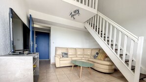 A living room with a beige sofa, a glass coffee table, and a TV on a stand. A staircase with a white railing is visible to the right. - Duplex T4 avec piscine, 6 couchages, garage, proche plage à Argelès-sur-Mer (Argelès-sur-Mer)