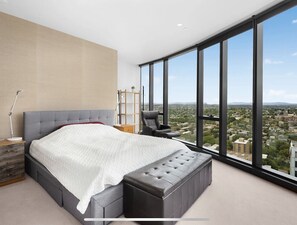 Relax in a spacious bedroom with a large bed, dark grey bench, and a tall bookshelf, offering a panoramic city view.