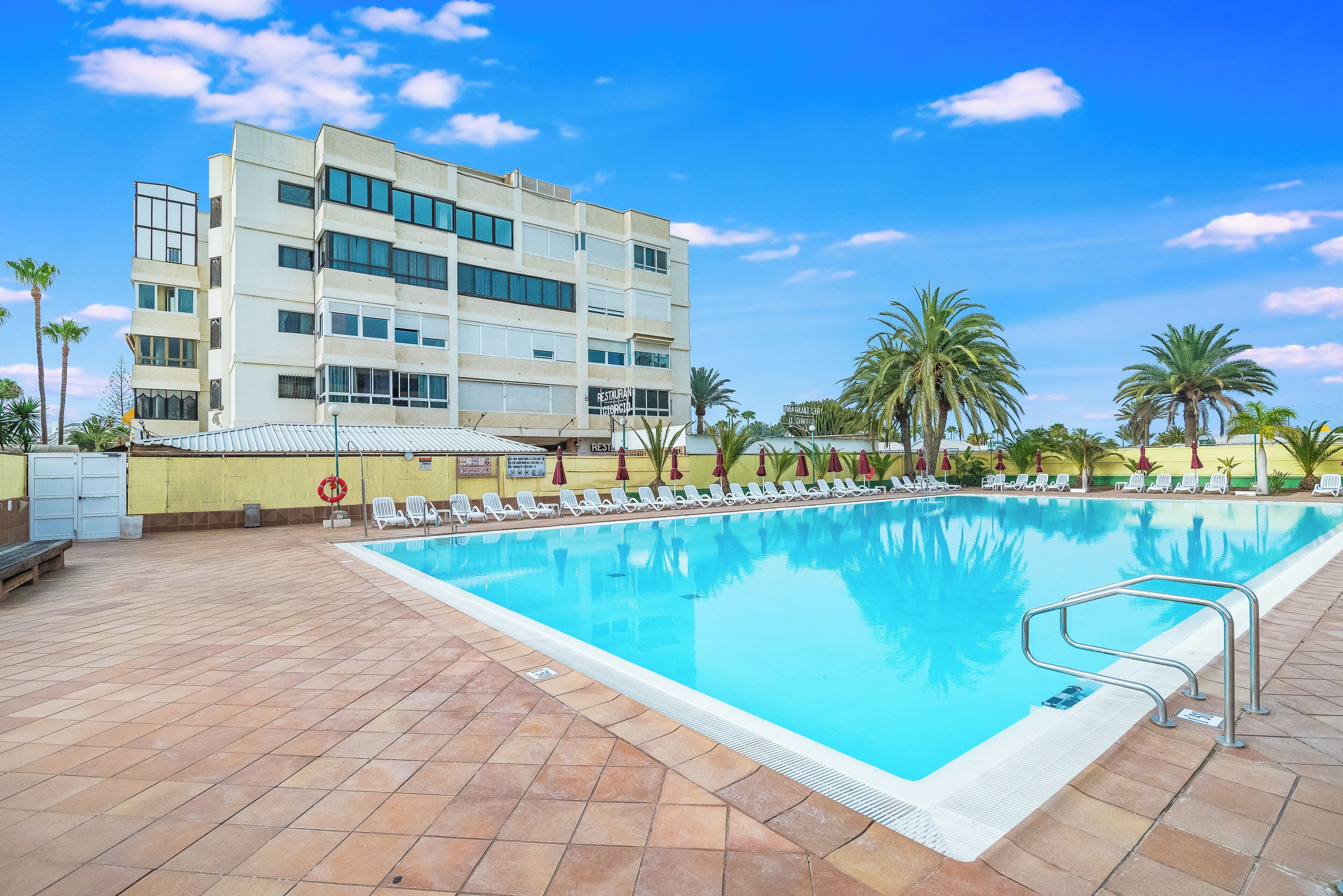 Una piscina con sillones de descanso y sombrillas, un edificio de varios pisos y palmeras.