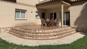 Un edificio de varios pisos con un patio cubierto que tiene una mesa, sillas, escalones de piedra y un pequeño ventanal con cortinas.