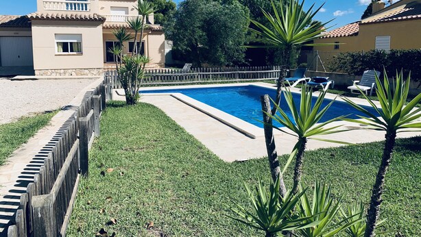 Una piscina rodeada por una valla de madera, con un área verde y un edificio de techo a tejas al fondo.