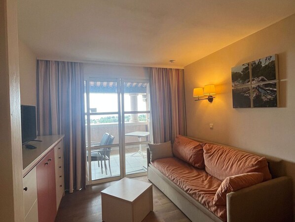 A room with a sofa, a small table, a TV on a cabinet, a sliding glass door leading to an outdoor area, and a wall-mounted lamp. - 3-room duplex with solarium and pool access (Saint-Raphaël)