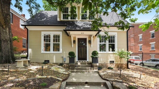 A two-story house with a beige exterior, numbered 614, featuring a front porch with steps leading to the door, potted plants, and a stone pathway.