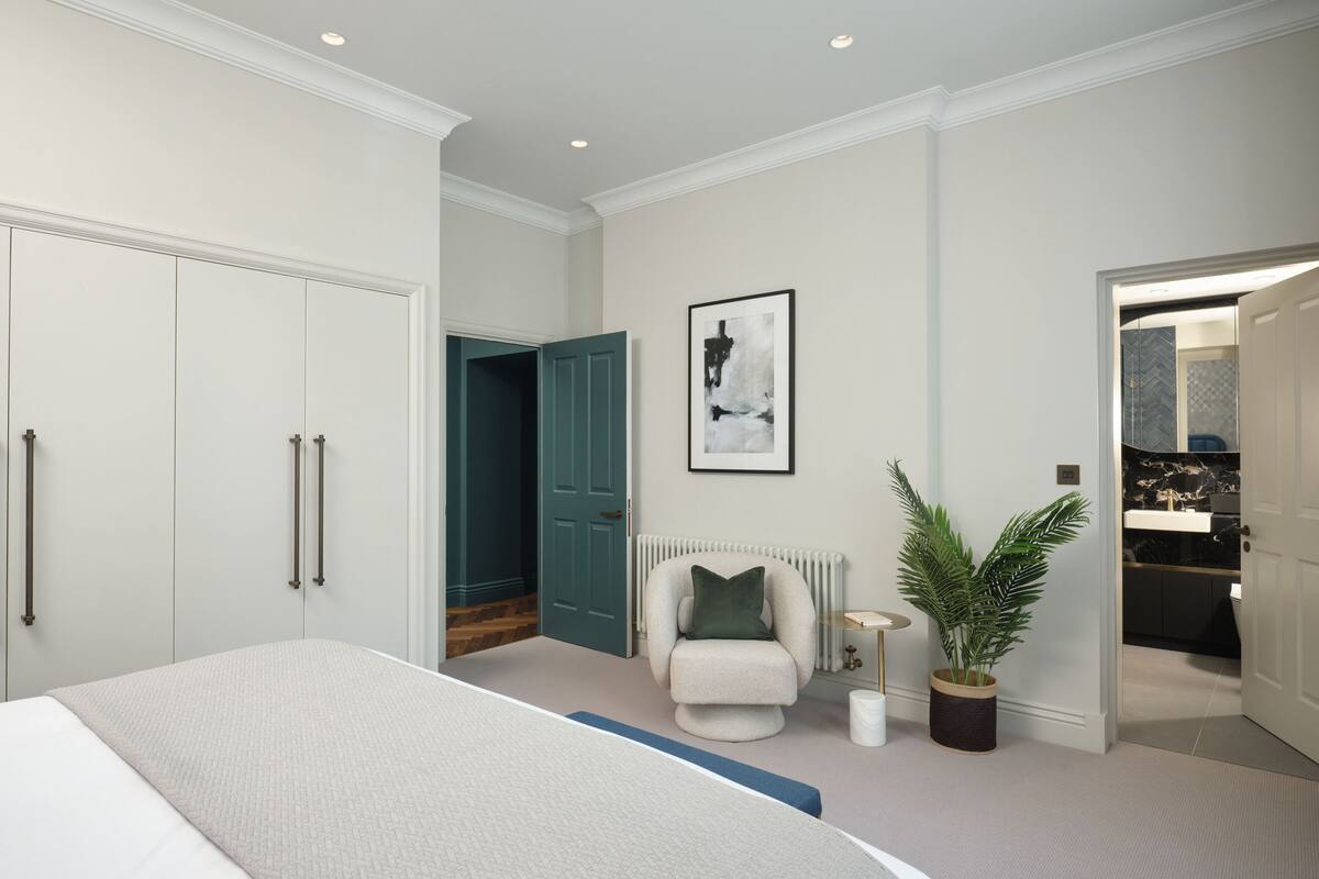 A bedroom with a bed, a chair, a plant, and a bathroom visible through an open door.
