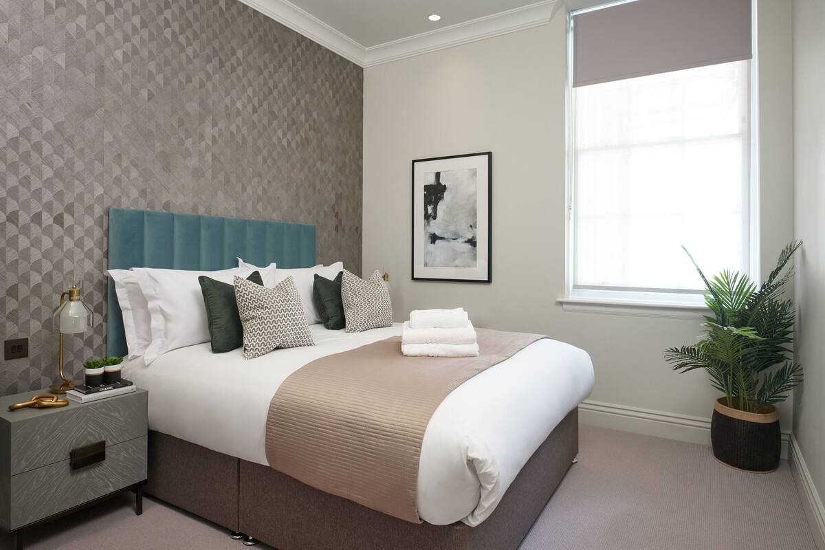 A modern bedroom with a large bed, a nightstand, a potted plant, and a window with blinds.