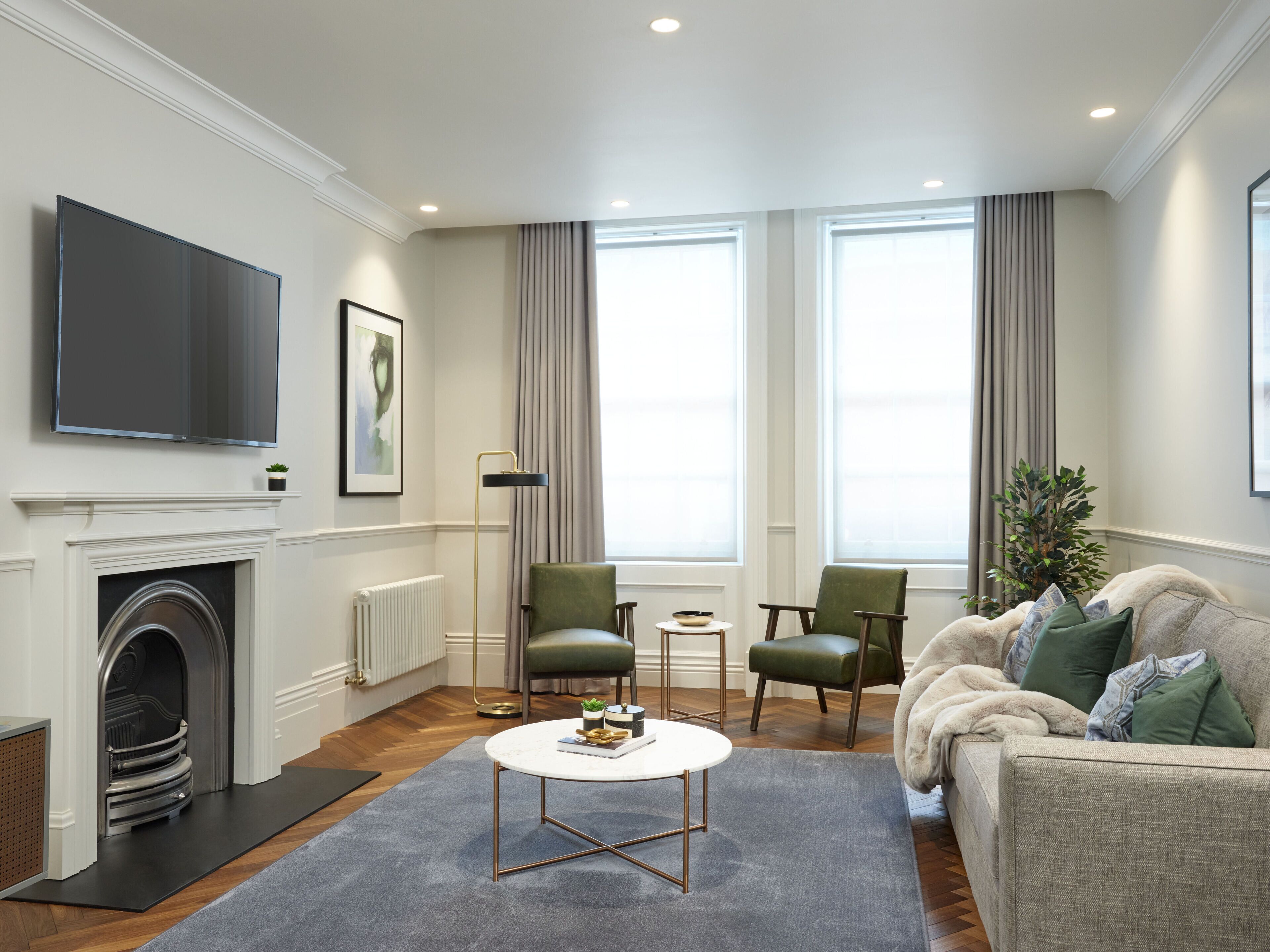A modern living room with a fireplace, a flat-screen TV, two green armchairs, a white coffee table, and a grey sofa with green cushions.
