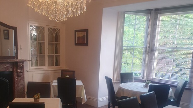 A dining area with tables and chairs, a chandelier, a window with blinds, and a cabinet with glass doors. - The Shrubbery Hotel (Shepton Mallet)