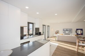 Eine moderne Küche mit einer großen Kochinsel, ein Essbereich mit Holztisch und -stühlen sowie ein Wohnzimmer mit weißem Sofa und Fernseher.