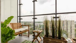 A modern room with a large window, a wooden bench, a small table, and potted plants. - Stylish Studio in Wola | Air Conditioning and Balcony (Warszawa)