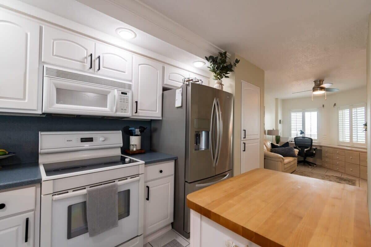A modern kitchen with white cabinetry, a stainless steel refrigerator, a built-in microwave, and a wooden countertop.