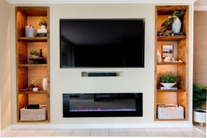 Un salon moderne avec un grand téléviseur à écran plat fixé à un mur blanc, de part et d’autre duquel se trouvent des étagères en bois ornées d’objets décoratifs.