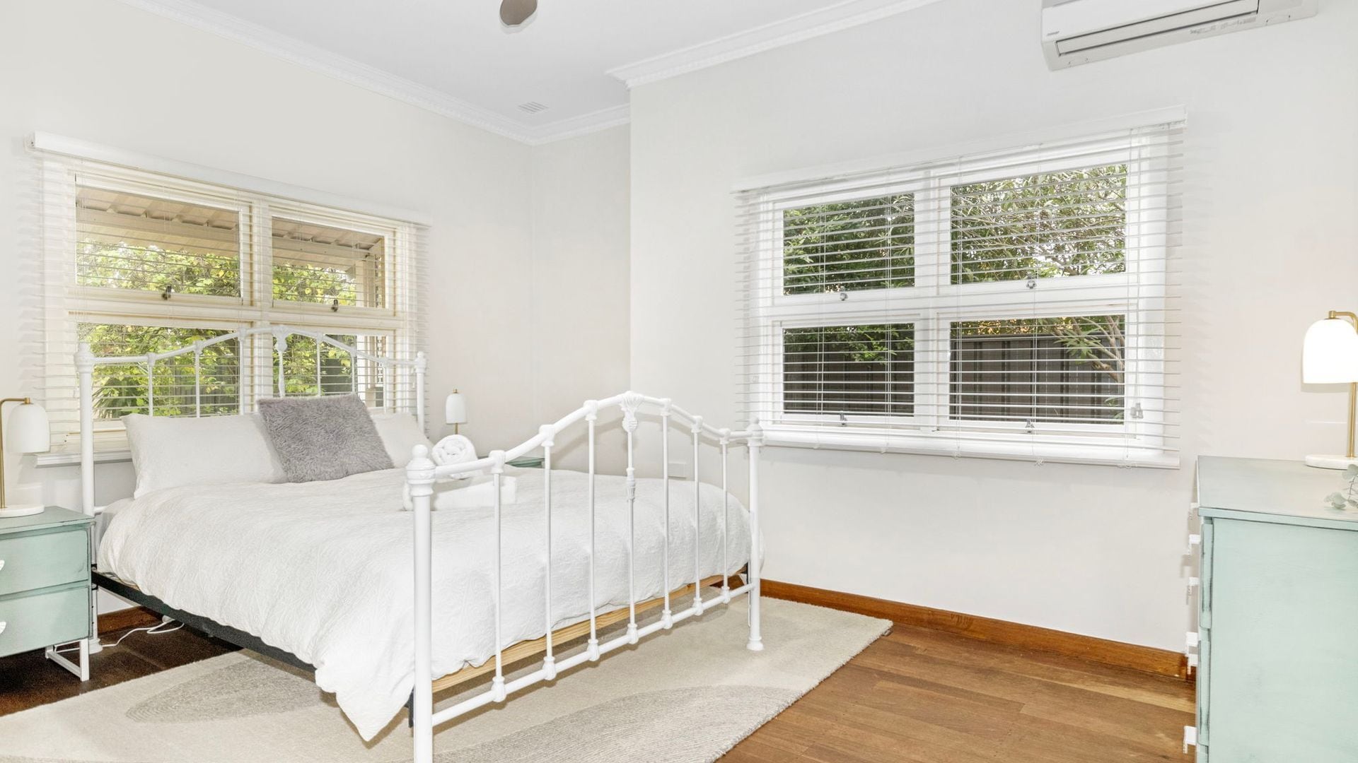 A bedroom with a white metal bed frame, a white bedside table, a nightstand, a window with blinds, and a wall-mounted air conditioning unit.