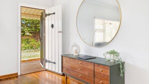 Una habitación con piso de madera, una puerta blanca que da al exterior, un espejo redondo, una pequeña escultura blanca y un mueble de madera oscura con cajones.