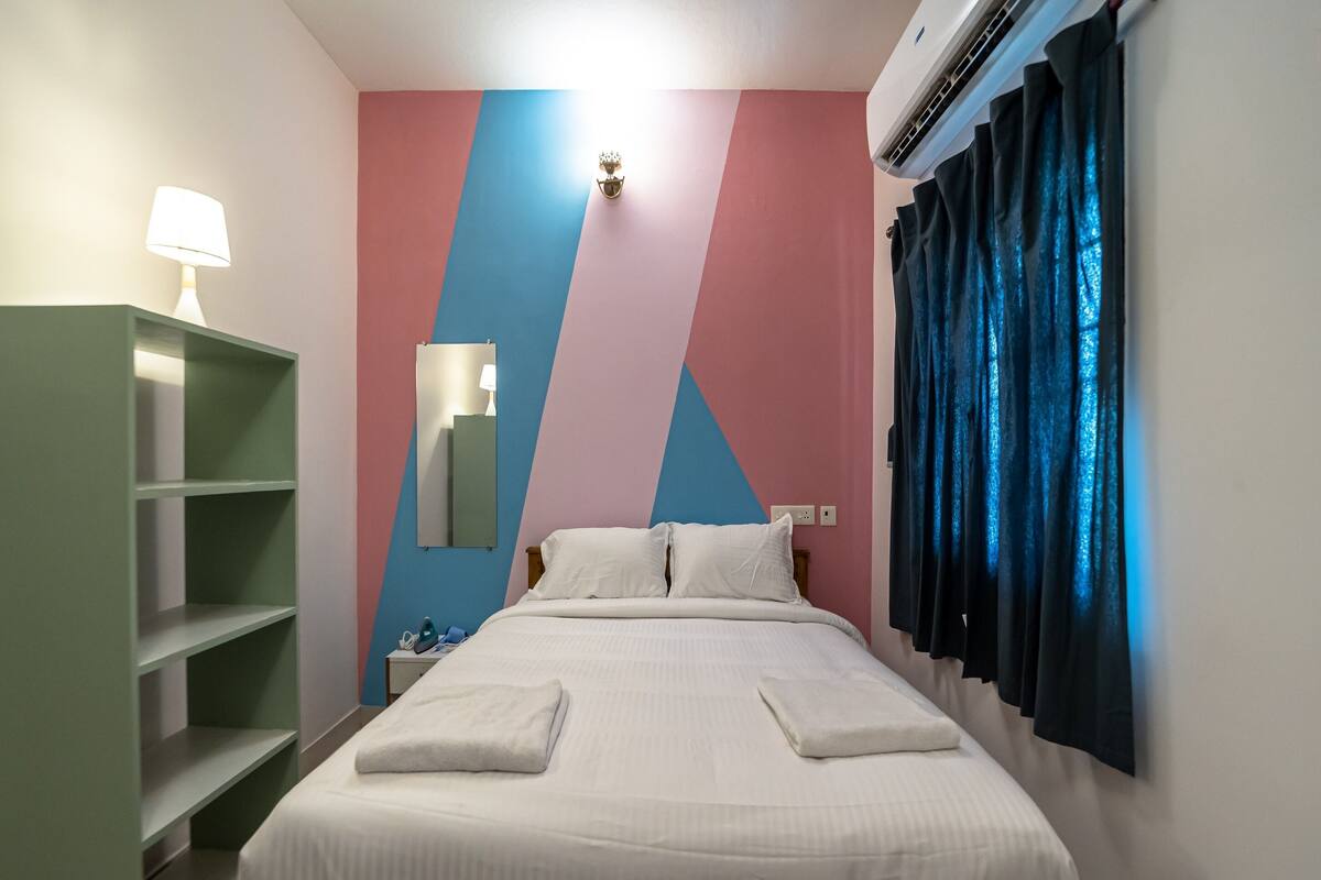 Una cama bien hecha con almohadas, un armario verde, un espejo fijado a la pared y una ventana con cortinas.