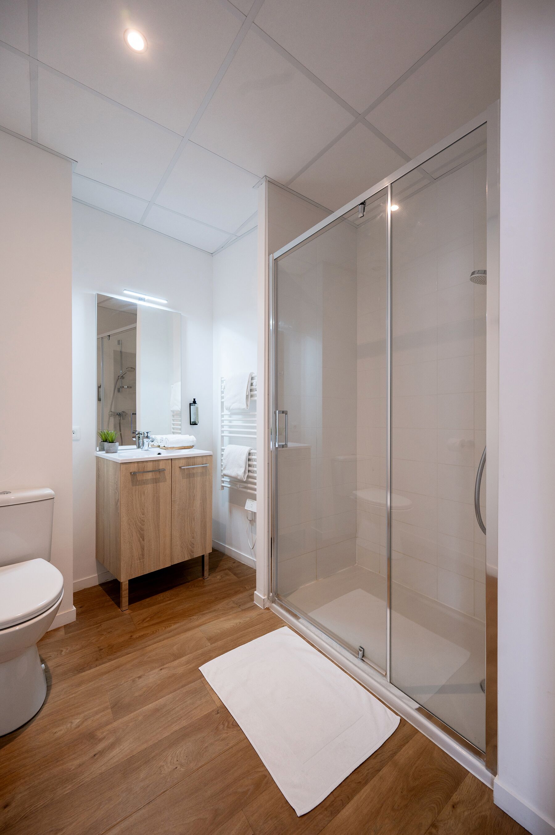 Ein modernes Badezimmer mit Toilette, Holzwaschtisch, Duschkabine aus Glas und einem weißen Teppich.