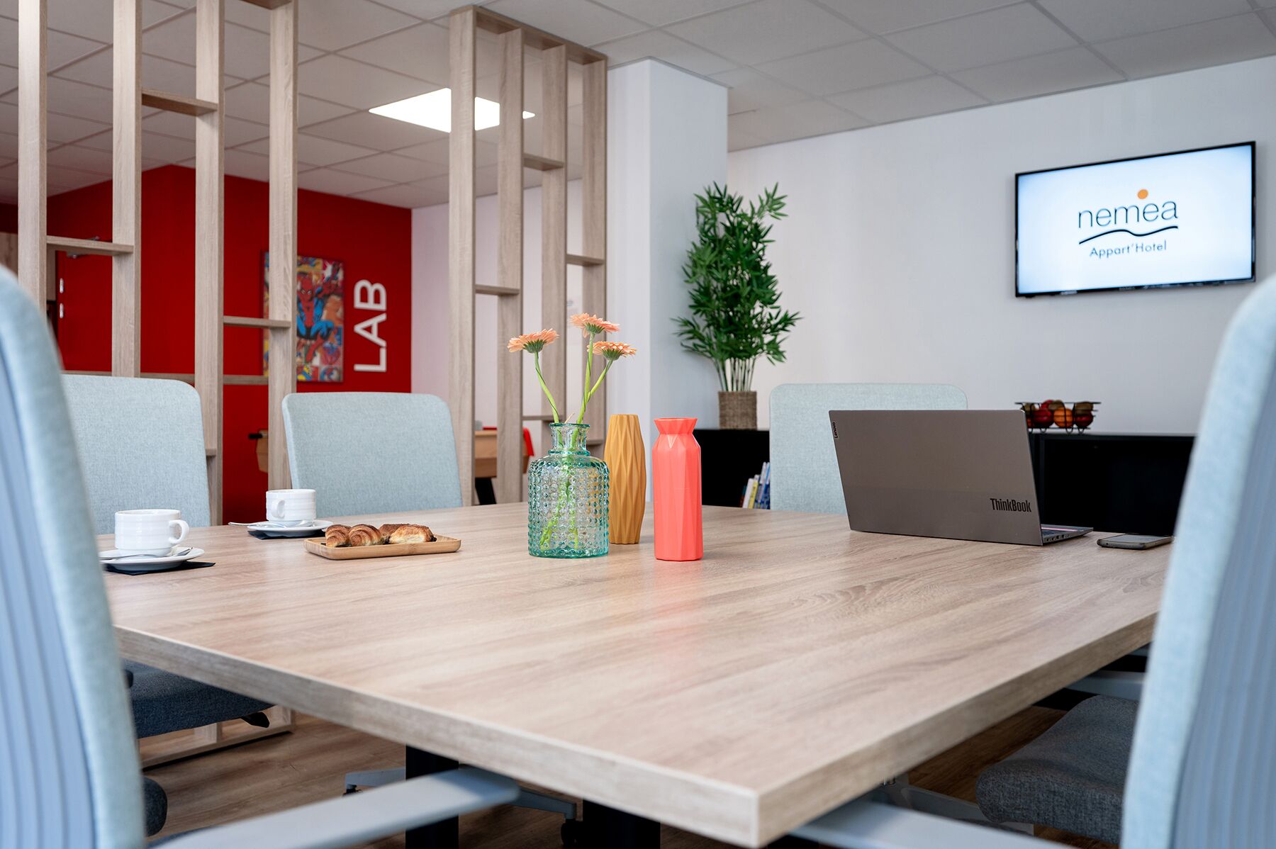 A conference room with a wooden table, a laptop, and a ThinkBook. There are chairs around the table, a plant, and a TV screen displaying 'nemea Appart/Hotel'.