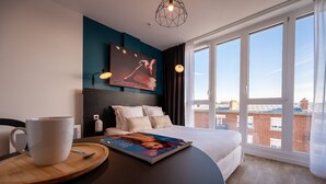A modern hotel room with a large bed, a dining table with a magazine, a coffee cup, and a city view through large windows. - Nemea Appart Hotel Coliseum Amiens Centre (Amiens)