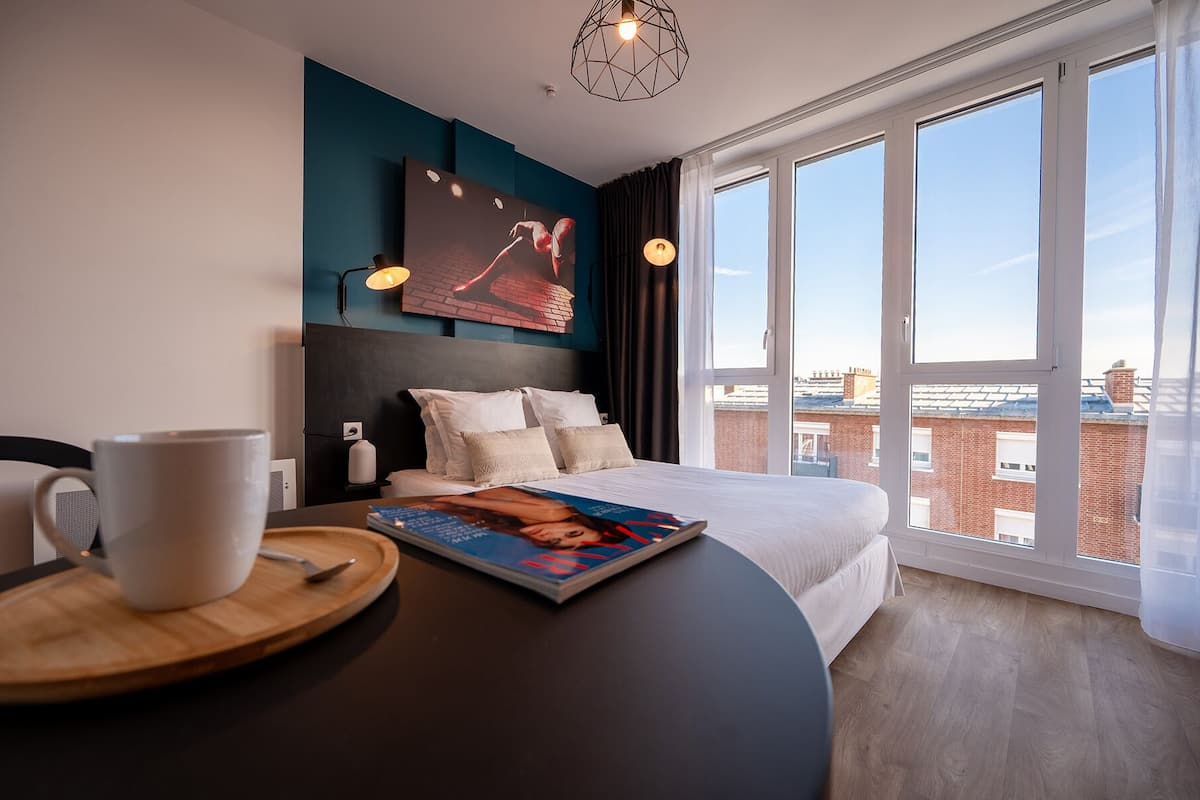 Ett modernt hotellrum med en stor säng, ett matbord med ett tidningsnummer, en kaffekopp och utsikt över staden genom stora fönster.