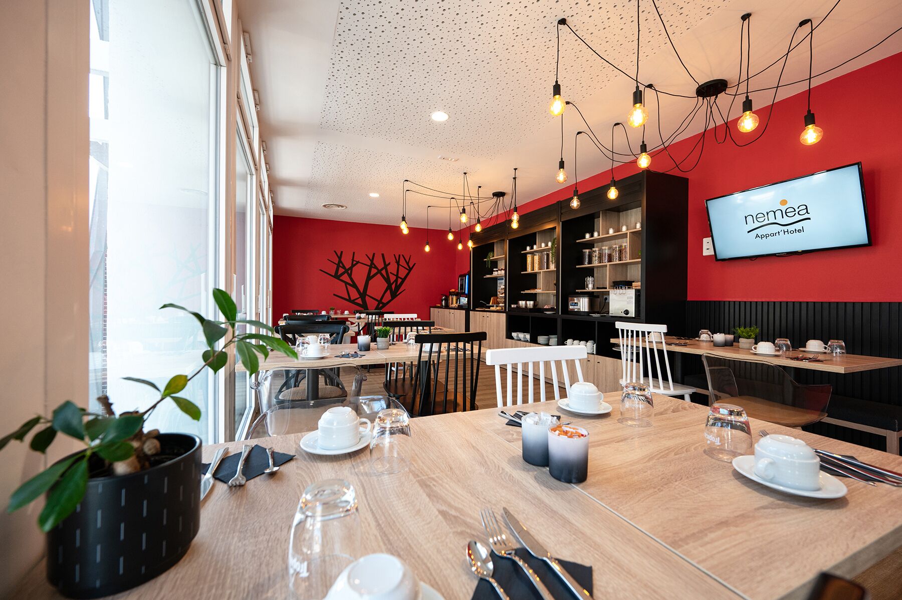 Un comedor moderno con pared roja, estanterías negras y un televisor mostrando 'nemea Appart'Hoteli'.