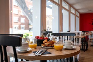 Una mesa de desayuno con cruasanes, manzanas y jugo de naranja en una habitación luminosa con ventanas grandes.