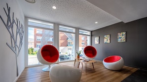 A modern interior with large windows, two red and white chairs, a wooden table, and a ceiling with a perforated pattern. - Nemea Appart Hotel Coliseum Amiens Centre (Amiens)