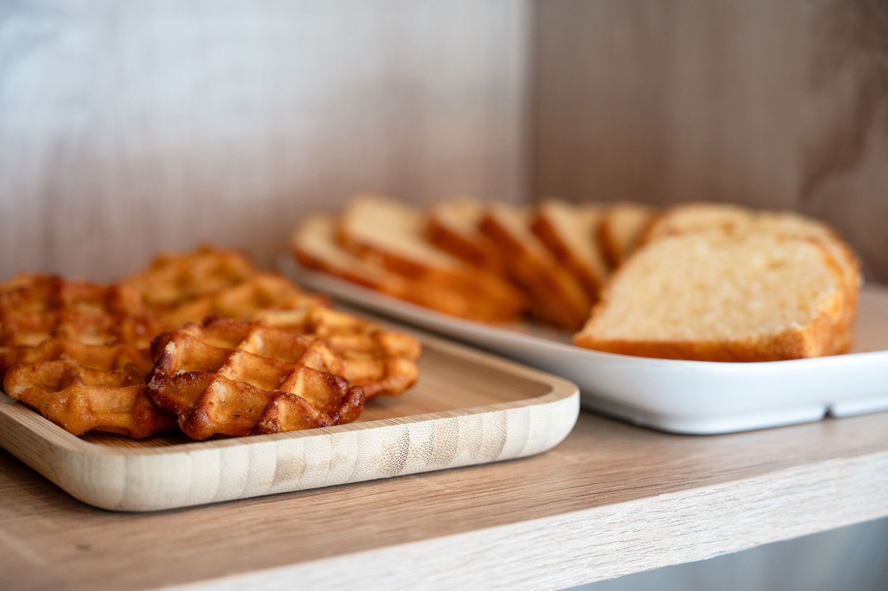 A wooden cutting board with waffles and a white plate with sliced bread.