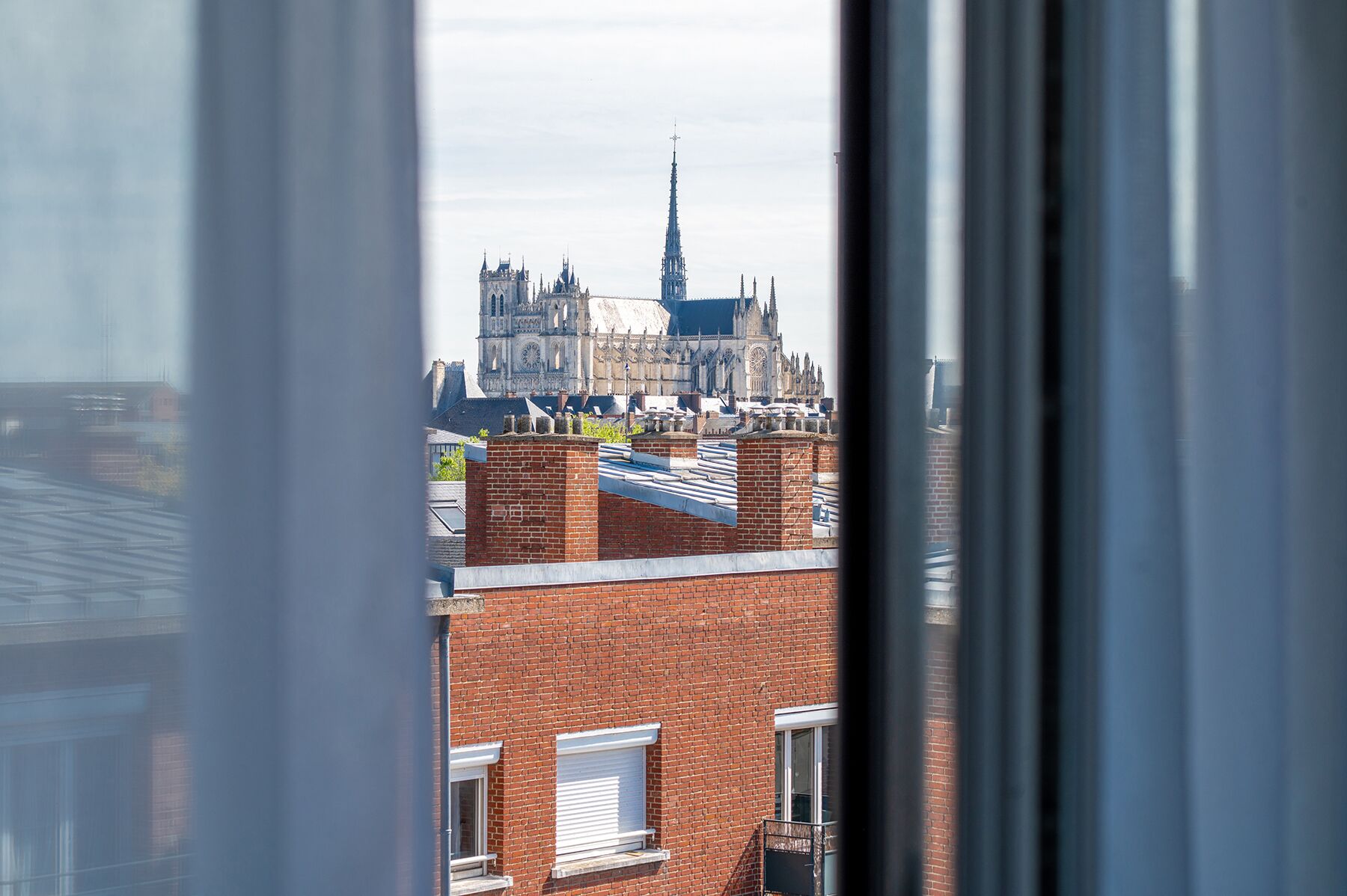 A view of a historic cathedral with intricate architecture and a tall spire, seen through a window with a brick building in the foreground.