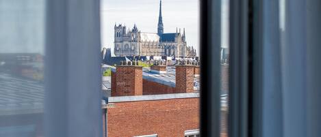 Une vue d’une cathédrale historique, caractérisée par son architecture complexe et son flèche imposante, que l’on aperçoit à travers une fenêtre, avec un bâtiment en briques en premier plan.