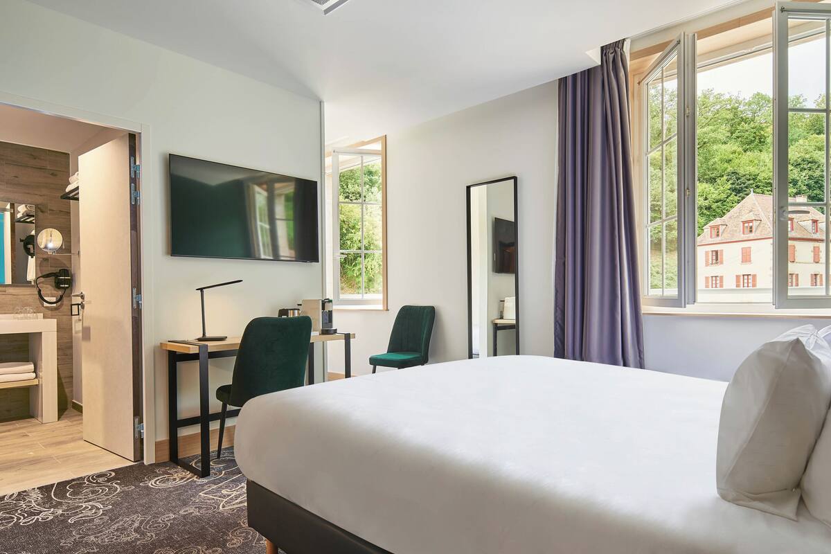 Habitación de hotel con una cama grande, un escritorio con silla, televisión y ventana con vistas a edificios.