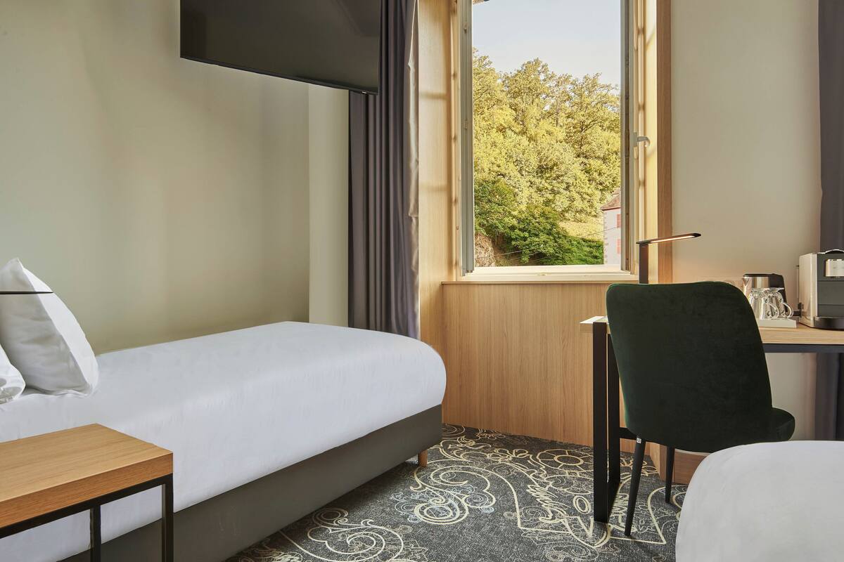 Habitación de hotel con cama, escritorio, silla, televisor montado en la pared y ventana con vista a árboles.