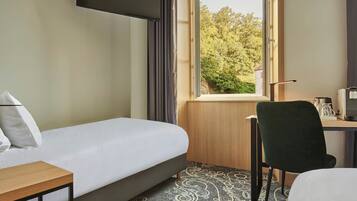 A hotel room with a bed, a desk, a chair, a TV mounted on the wall, and a window with a view of trees.
