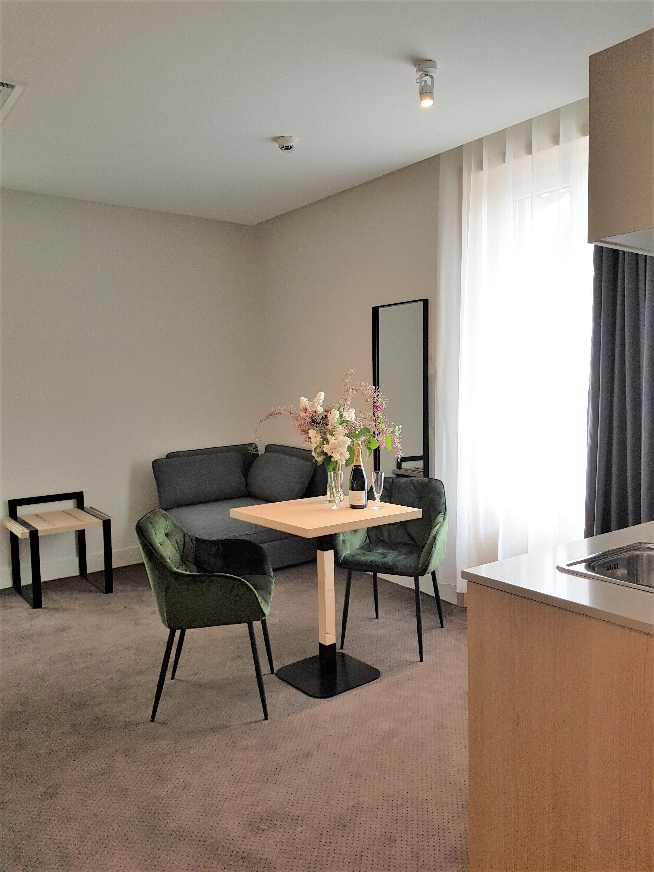 A modern hotel room with a sofa, two chairs, a small table with a vase of flowers, a mirror, and a kitchenette.