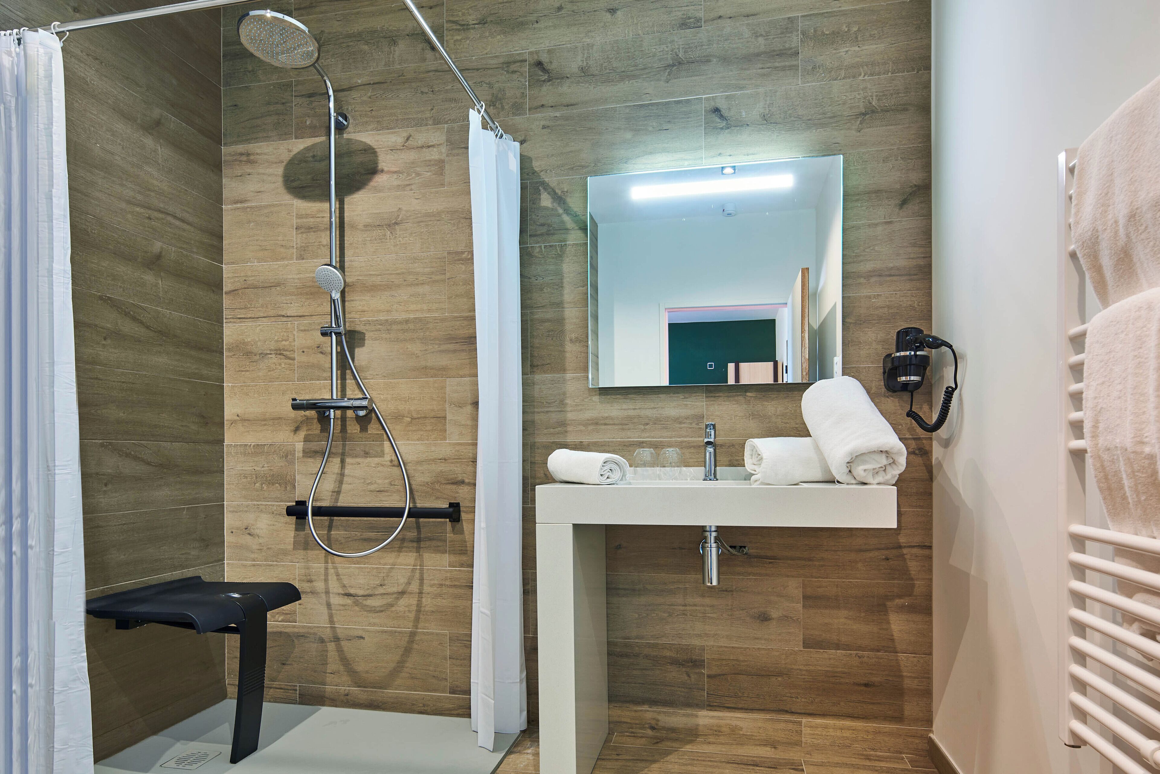 Une salle de bain moderne avec un plancher en bois, une douche équipée d’un pommeau de douche à main, un banc, un lavabo avec un miroir et un porte-serviettes.