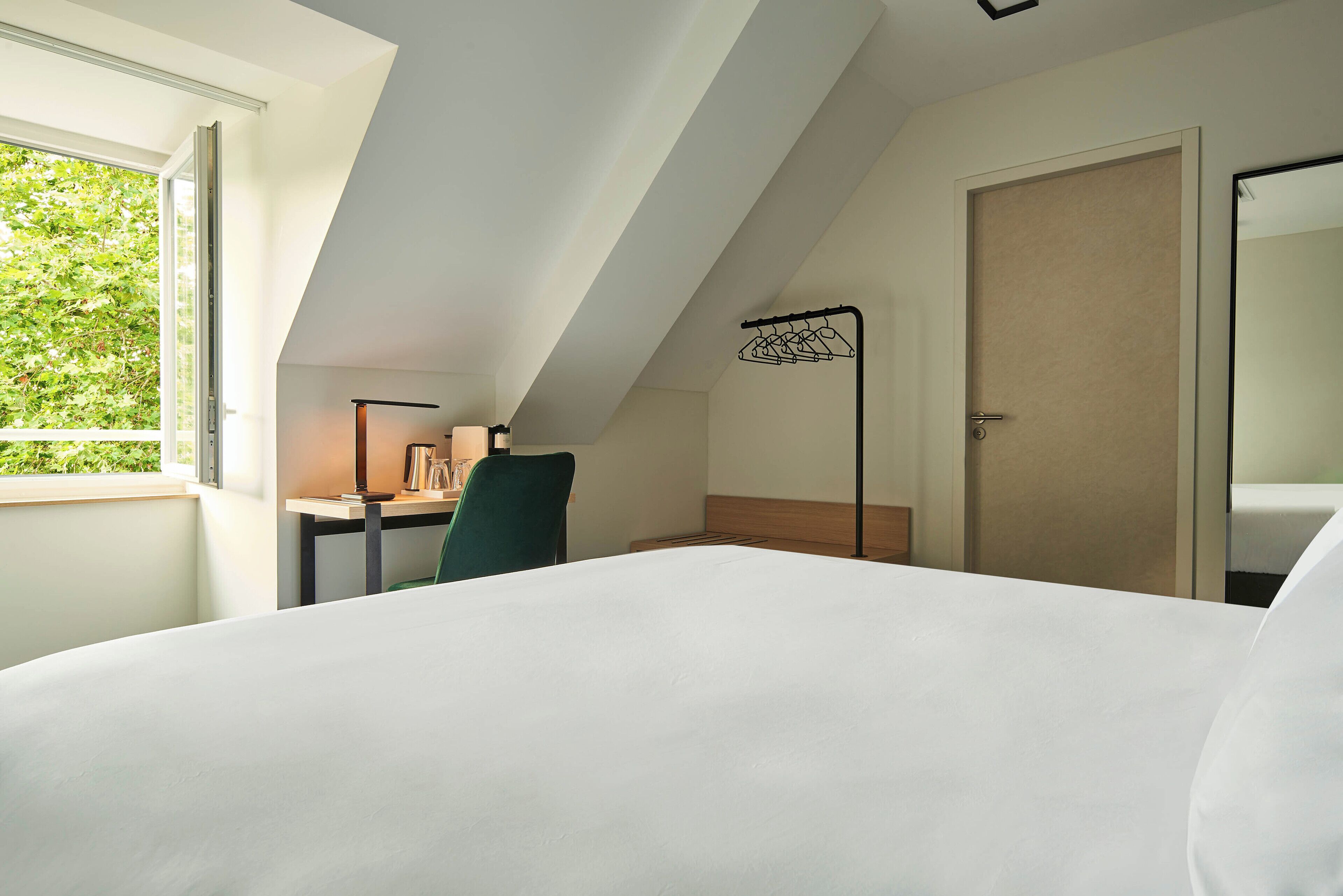 A hotel room with a large bed, a desk with a chair, a window with a view of greenery, and a door.