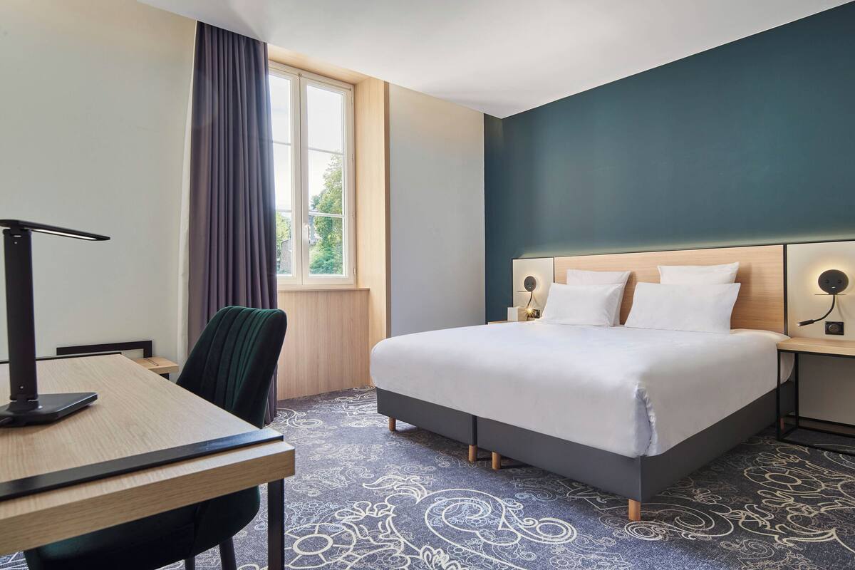 Una habitación de hotel con cama, escritorio, silla, ventana con cortinas y alfombra estampada.