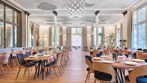 A spacious dining area with multiple tables and chairs, large windows, and a modern chandelier.