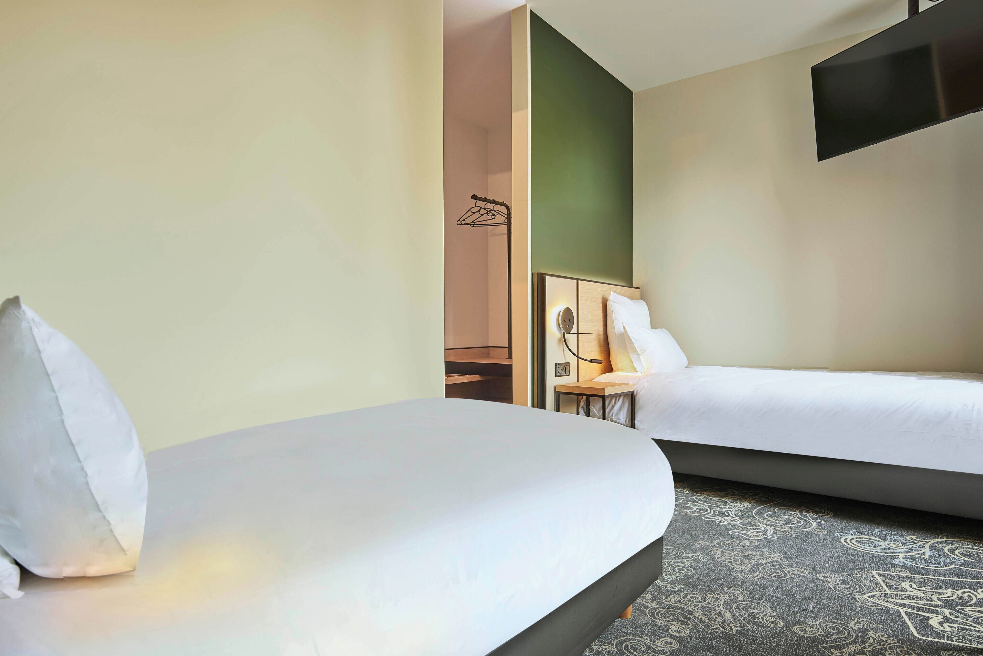 Una habitación de hotel con dos camas, un televisor montado en la pared y una mesita de noche con una lámpara.